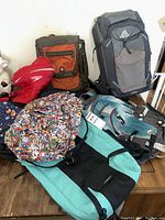 Photo shows six backpacks arranged on floor - Gregory gray backpack with blue accents, orange and gray Timbuk2 bag, red mesh Speedo swim bag, turquoise and black High Sierra backpack with tags, colorful Sherpani character pattern backpack, dark blue For U Designs space corgi print backpack.