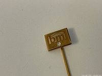 Close-up photo of the gold stick pin showing the rectangular face with 'bm' initials and tiny diamond below.