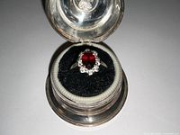 Ring displayed inside round silver-tone jewelry box on black velvet cushion, top-down view