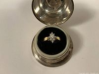 Ring displayed in round silver-tone hinged jewelry box, top-down view showing cluster diamond starburst design and yellow gold split shank.
