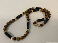 Full view of necklace laid flat showing alternating beads of round tiger eye stones, black rectangular beads, and ribbed 14k gold spacer beads.