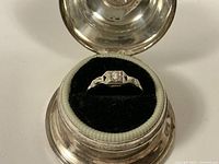 Ring in velvet-lined display box with hinged lid open, showing top view of bezel-set diamond