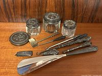 Full lot arrangement showing three cut glass powder jars topped with ornate sterling silver lids, an oval silver compact, two ornate silver handled nail/tool implements, shoehorn, buttonhook, and other small grooming tools.