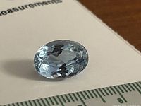 Close-up image of an oval-cut light blue topaz gemstone on a white paper background with a ruler for size reference.