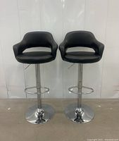 Pair of modern black leather-like barstools front view showing seats with curved backs and armrests, chrome pedestal bases with footrests.