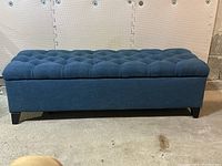 Front view of the blue fabric storage bench showing the tufted cushioned lid and dark tapered legs on a plain background.