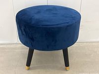 Royal blue velvet round ottoman with black legs and gold-tone caps shown from front angle on concrete floor against white wall.