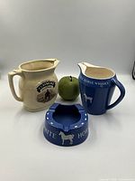 Front view of the three items: cream Claymore whiskey water pitcher, blue White Horse Whisky pitcher, and blue White Horse ashtray with white horse emblem, with a green apple for scale.