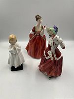 Three Royal Doulton porcelain figurines shown together: two women in period costume and one child in white gown.