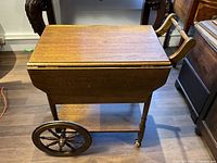 Full view of the antique wooden tea trolly or bar cart with drop leaves closed, extended handle and spoked wooden wheels with rubber tread.