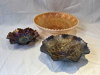 Three art glass serving pieces displayed on white cloth, showing an orange bowl and two iridescent ruffled bowls