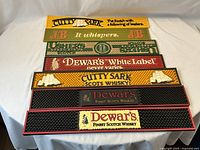 All 7 rubber bar mats laid out on a white table showing various whisky brand logos and slogans.