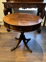 Photo showing full round mahogany drum table with pedestal base and single drawer, placed on wood floor with another carved wood table in background.
