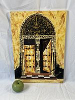 Oil painting showing front view of arched doorway with doors open, yellow ochre background, intricate patterns on arch and door frame, with green apple next to painting for size reference.