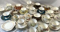 Photo showing full lot of 18 tea cups laid with their matching saucers, diverse styles and patterns, predominantly floral with gold accents, white and colored porcelain arranged in rows.