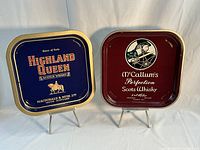Both vintage Scotch whiskey serving trays displayed side by side on stands, Highland Queen tray blue with gold trim, McCallum's tray maroon with artwork.