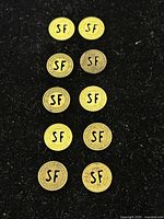 Top-down view of 10 vintage San Francisco municipal fare transportation tokens arranged in two columns on a black textured surface. Tokens exhibit various designs with 'SF' initials and circular inscriptions.