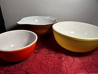 Three vintage Pyrex glass mixing bowls shown upright. From left to right: small orange bowl, large brown bowl with pouring spout, medium yellow bowl.