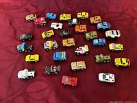 Top view of 30 assorted Micro Machines vehicles in various colors arranged on a red fabric background showing the variety and general condition of each vehicle.