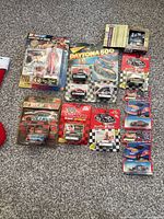 Lot of nine unopened NASCAR diecast cars and figures arranged on carpet showing packaging and size variety.