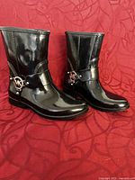 Front and side views of shiny black Michael Kors women's short rain boots, size 7, showing MK logo and glossy finish on a patterned red background.