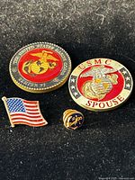 Photo of two US Marine Corps coins, one with Semper Fi text and another USMC Spouse coin, alongside an American flag pin and a small USMC emblem pin.