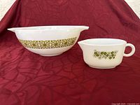 Pair of white Pyrex items: a 10 inch mixing bowl decorated with an olive green geometric floral band and a gravy boat with olive green floral decoration, set against a red patterned fabric background.
