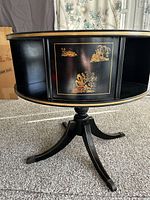 Side view showing black lacquered finish, gold chinoiserie artwork on rotating panel, pedestal base with four legs.