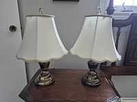 Pair of bronze bedside lamps with white scalloped fabric shades on a wooden surface, front view showing shape and color.