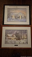 Two framed winter prints stacked vertically on a wooden floor, one with a horse-drawn sleigh and another showing a snowy house with trees and a fence.