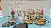 Photo showing 14 assorted candle holders including oil lamps with glass chimneys and smaller brass and glass candle holders of various styles and sizes arranged on a blue textured surface.