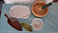 Clear glass rectangular baking dish, white oval ceramic dish, wooden salad bowl with salad utensils, small glass dome, round ceramic bowl with floral pattern, and two leaf-shaped ceramic dishes.