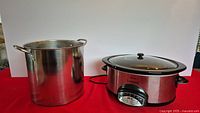 Rival Crock-Pot slow cooker and large stainless steel chili pot side by side on a red surface against a plain background