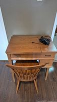 Wooden sewing table closed with matching chair in front. Visible light wear and scratches on wood surface. Power cord coiled on top of table.