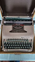 Top down view of the Smith-Corona Sterling typewriter showing greenish-gray body, round teal keys, ribbon spool, and partial view of carrying case interior.