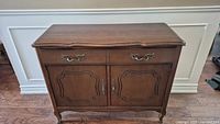 Front view of a traditional French Provincial brown wooden sideboard with two drawers and two-door cabinet. Ornate metal handles and carved drawer/cabinet fronts. Shows scuffs and wear on surface and legs.