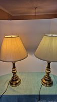 Two traditional style table lamps with polished metal bases and pleated lampshades on table with blue cloth backdrop. Lamps are illuminated.