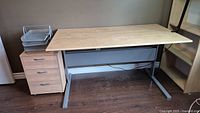 Desk with light wood finish and silver metal legs, accompanied by a filing cabinet with three drawers.
