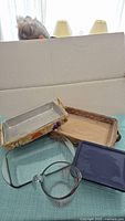 Photo showing two woven baskets, one containing a metal dish, the other a glass dish, a clear glass 4-cup measuring cup, a blue lidded rectangular container, and glass Pyrex bakeware pieces underneath.