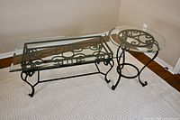 Both coffee and side tables shown together on light carpet, highlighting black wrought iron bases and clear glass tops.