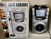 Karaoke machine next to its original box showing IXT CD+G Karaoke model GM655, with a 7-inch TFT color display, speaker, controls, and microphones attached.