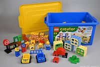 Set of Duplo pieces including figures, stop sign, windows, doors, fences, cars and printed bricks arranged in front of a blue Lego Creator storage box with yellow lid.