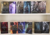 Photo of all 12 books arranged in two rows on a white shelf, showing front covers with fantasy-themed artwork and titles by Larissa Ione, Sherrilyn Kenyon, and Jacquelyn Frank.