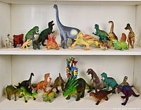 Two white shelves display a total of 57 dinosaur toys including 27 medium-large and 30+ small size figures. Top shelf has larger dinosaurs including long-necked, horned, and armored types. Bottom shelf has smaller to medium size dinosaurs with diverse species and bright colors.