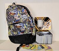 Full view of Marvel comic style backpack next to Pokemon tin with its lid open showing a large stack of Pokemon cards inside as well as some cards scattered in front.