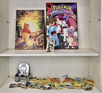 Full view of lot showing Winnie the Pooh poster on board, Pokemon poster on board, used gold Pokemon wallet, Pokemon tin and collectible cards spread around.