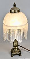 Lamp lit showcasing mottled frosted glass shade and beaded fringe with brass-colored base on neutral background.
