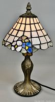 Full view of vintage Tiffany style mini table lamp with cream grid-patterned stained glass shade and floral accents in pastel colors. Ornate sculpted bronze-tone base with cord attached.