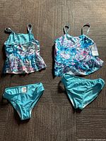 Two girls two-piece swimsuits laid flat showing tops with blue, purple, and white pattern and turquoise bottoms, one new with tags