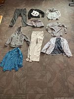 Overview of nine clothing items laid out on carpet including pants, sweaters, skirt, blouse, and blazer
